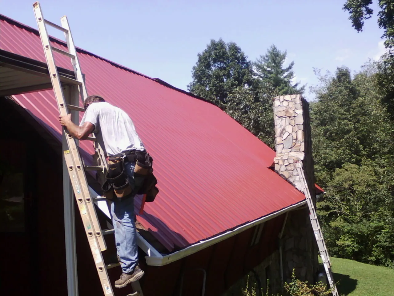 Roofer on a ladder beside a red metal roof and stone chimney — Soffit & Fascia Repair by Roofing Brothers in New Pine Creek